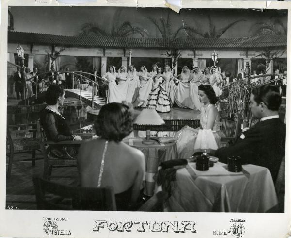 Scena del film "Fortuna" - Neufeld, Max, 1940 - Al centro del palco, Jone Salinas. Dietro di lei, dodici attrici non identificate con la mano destra che tiene un lembo della lunga gonna bianca. In primo piano, Maria Denis seduta.
