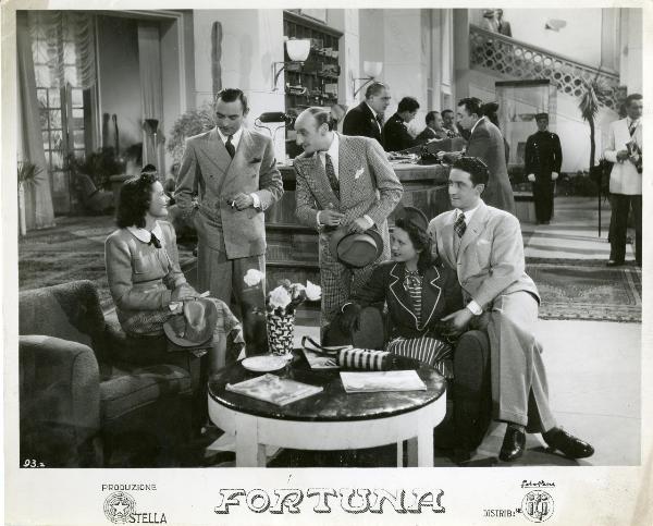 Scena del film "Fortuna" - Neufeld, Max, 1940 - Nella hall di un Hotel, Maria Denis, a sinistra, e Jone Salinas, a destra, sedute su dei divanetti parlano con, da sinistra a destra: Tony D'Algy, Manuel Paris e Luis Prendes.
