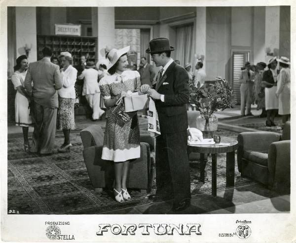 Scena del film "Fortuna" - Neufeld, Max, 1940 - Nella hall di un Hotel: Maria Denis e Tony D'Algy si guardano sorridendo mentre le loro mani si sfiorano tenendo in mano una borsa. Intorno a loro, attori non identificati.
