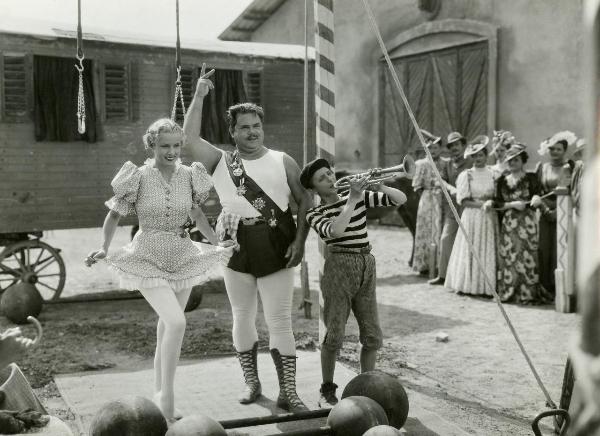 Scena del film "La forza bruta" - Bragaglia, Carlo Ludovico, 1941 - Juan De Landa, al centro, con il braccio sollevato. Maria Mercader, a sinistra, si solleva la gonna con entrambe le mani in un inchino e Sergio Tedesco, a destra, suona una tromba.