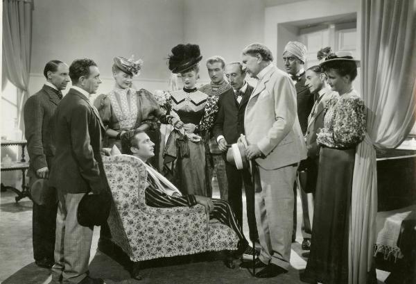 Scena del film "La forza bruta" - Bragaglia, Carlo Ludovico, 1941 - Rossano Brazzi, seduto sulla poltrona, è circondato da attori e attrici non identificati che lo guardano.

