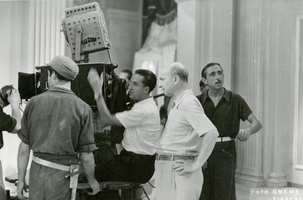 Sul set del film "Fra Diavolo" - Zampa, Luigi, 1942 - Giuseppe Guarino, con le mani sui fianchi, alle spalle di Luigi Zampa mentre gesticola guardando nella macchina da presa. Intorno a loro, altri operatori non identificati.
