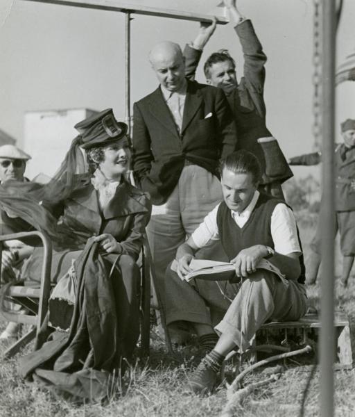 Sul set del film "Fra Diavolo" - Zampa, Luigi, 1942 - Laura Nucci, a sinistra seduta, guarda verso destra. Di fianco seduto, il regista Luigi Zampa legge dei fogli. Dietro, Giuseppe Guarino li osserva. Attorno, altri operatori non identificati.
