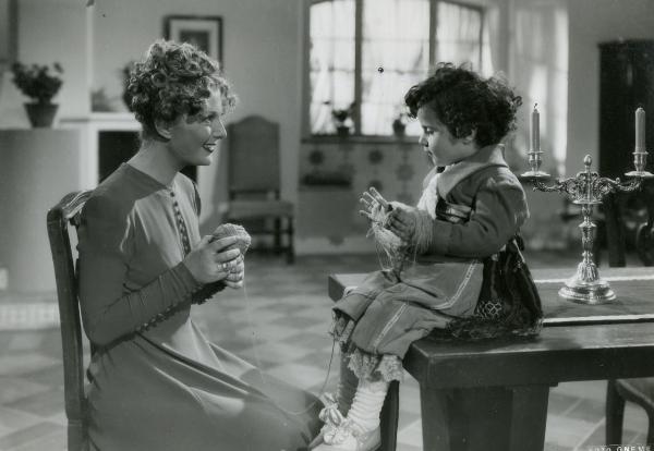 Scena del film "Fra Diavolo" - Zampa, Luigi, 1942 - Laura Nucci, seduta a sinistra, tiene tra le mani un gomitolo di tessuto mentre, sorridendo, guarda la bambina seduta sul tavolo di fronte a lei che tiene tra le mani il filo.

