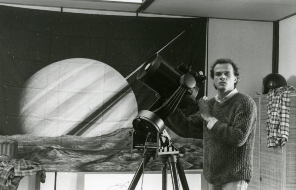 Scena del film "Francesca è mia" - Russo, Roberto, 1986 - Pierre Malet guarda verso la telecamera mentre sta utilizzando un telescopio. Sullo sfondo, un telo raffigurante Saturno.
