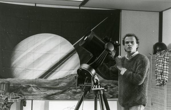 Scena del film "Francesca è mia" - Russo, Roberto, 1986 - Pierre Malet guarda verso la telecamera mentre sta utilizzando un telescopio. Sullo sfondo, un telo raffigurante Saturno.
