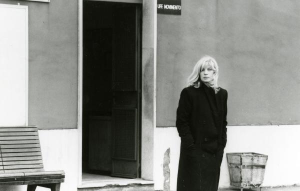 Scena del film "Francesca è mia" - Russo, Roberto, 1986 - Monica Vitti, in piedi, porta un lungo cappotto e tiene le mani in tasca. Dietro di lei, la porta aperta di un ufficio, un cestino a destra e una panchina a sinistra.
