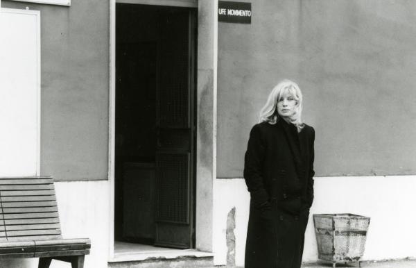 Scena del film "Francesca è mia" - Russo, Roberto, 1986 - Monica Vitti, in piedi, porta un lungo cappotto e tiene le mani in tasca. Dietro di lei, la porta aperta di un ufficio, un cestino a destra e una panchina a sinistra.
