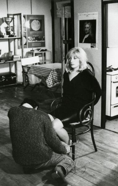 Scena del film "Francesca è mia" - Russo, Roberto, 1986 - Monica Vitti, seduta al centro della stanza, guarda dritto davanti a sé. Un attore non identificato di spalle le sta legando i piedi con una corda alla sedia.
