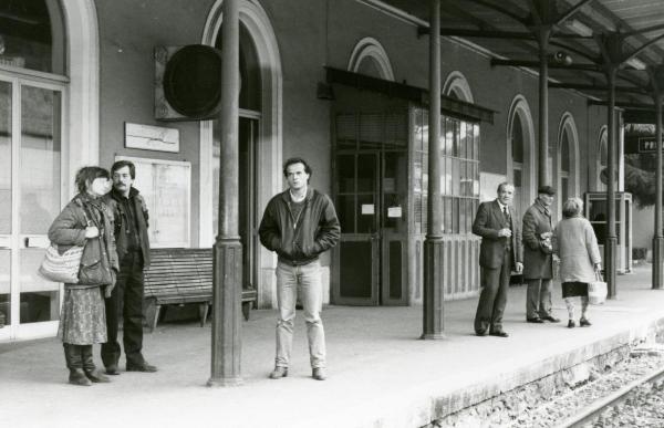 Scena del film "Francesca è mia" - Russo, Roberto, 1986 - Pierre Malet fermo in piedi sul binario della stazione che con le mani nelle tasche della giacca di pelle. Intorno a lui, attori non identificati aspettano il treno.
