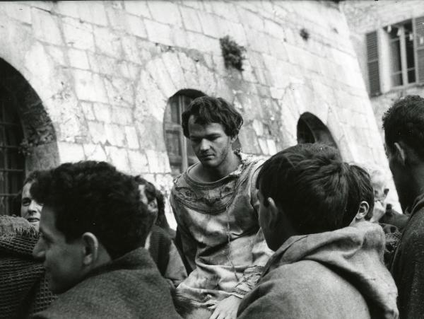 Scena del film "Francesco d'Assisi" - Cavani, Liliana, 1966 - Lou Castel, al centro, in vesti da Francesco d'Assisi, guarda in basso. Attorno a lui, in primo piano, attori non identificati di spalle.
