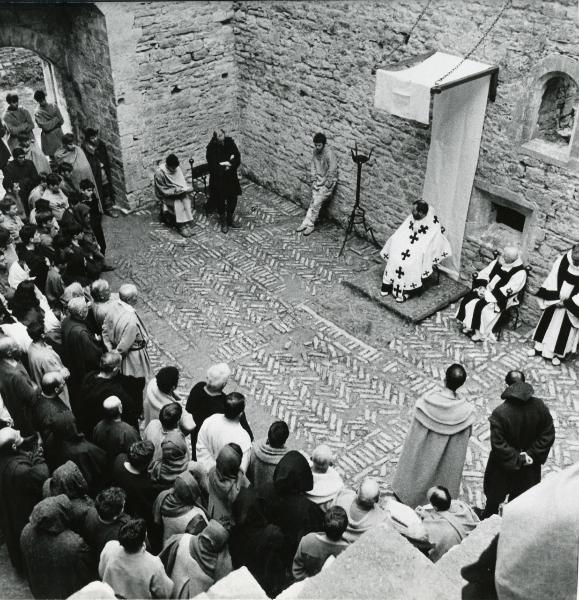 Scena del film "Francesco d'Assisi" - Cavani, Liliana, 1966 - Sotto un baldacchino, un attore non identificato in veste sacerdotale guarda verso Lou Castel, a sinistra, appoggiato a un muro. Di fronte a loro, una folla di persone li guarda.