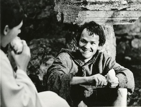 Scena del film "Francesco d'Assisi" - Cavani, Liliana, 1966 - Al centro, Lou Castel, con in mano un tozzo di pane, guarda sorridente verso sinistra un attore non identificato mentre sta mangiando.
