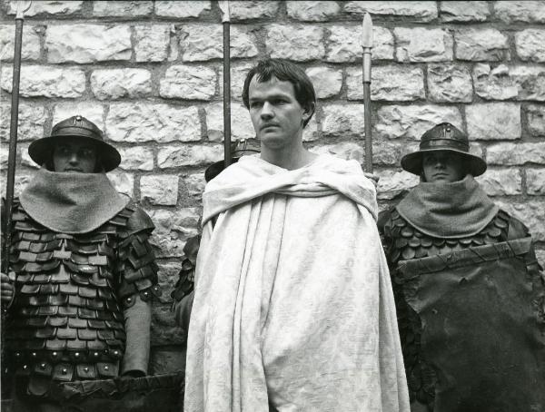 Scena del film "Francesco d'Assisi" - Cavani, Liliana, 1966 - Al centro, Lou Castel, vestito con un soprabito damascato, guarda verso sinistra. Dietro di lui, tre attori non identificati nei panni di guardie.