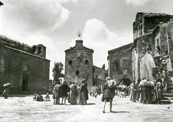 Scena del film "Francesco, giullare di Dio" - Rossellini, Roberto, 1950 - Al centro di una piazza, molti attori non identificati sono ritratti durante la loro vita quotidiana.