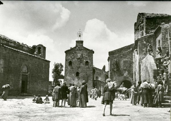 Scena del film "Francesco, giullare di Dio" - Rossellini, Roberto, 1950 - Al centro di una piazza, molti attori non identificati sono ritratti durante la loro vita quotidiana.