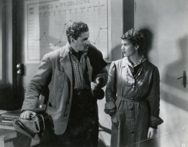 Scena del film "I fratelli Castiglioni" - D'Errico, Corrado, 1939 - Luisa Ferida in impermeabile a destra, guarda Amedeo Nazzari a sinistra che le parla gesticolando con la mano sinistra. Dietro di essi una lavagna con disegnato un grafico.