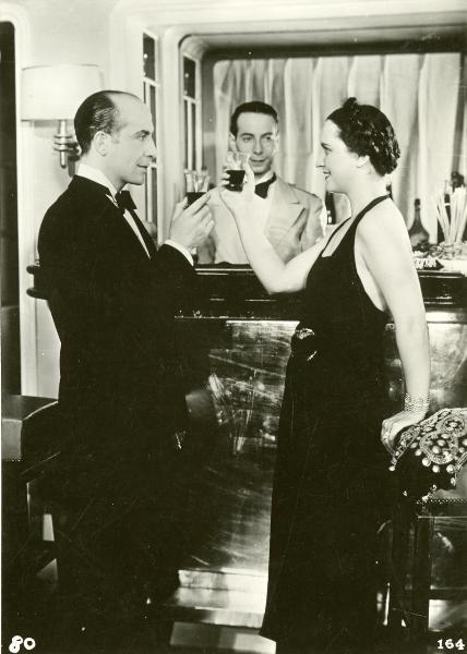 Scena del film "Freccia d'oro" - D'Errico, Corrado, 1935 - In primo piano, un attore non identificato e Vanna Vanni brindano guardandosi negli occhi. Dietro, un attore non identificato nei panni di cameriere li guarda.