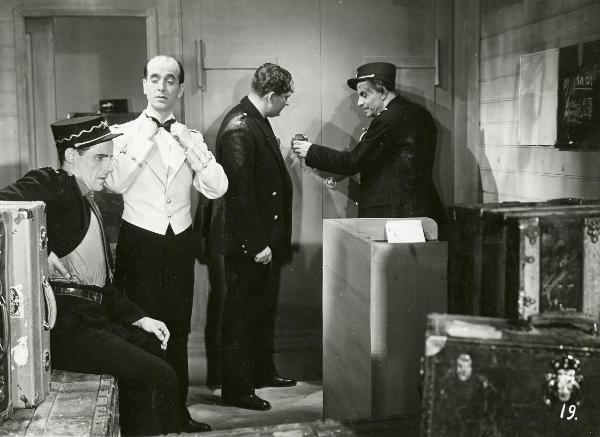 Scena del film "Freccia d'oro" - D'Errico, Corrado, 1935 - Quattro attori non identificati: il primo a sinistra seduto su un baule si tocca con la schiena, il secondo sistema il papillon. Gli altri due cercano di scassinare una cassaforte.