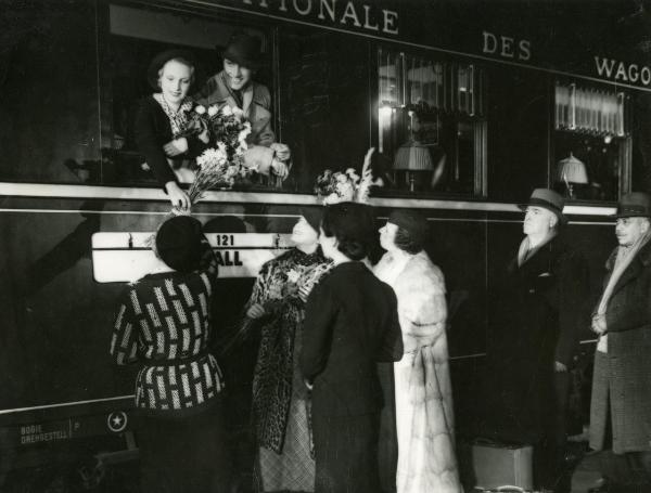 Scena del film "Freccia d'oro" - D'Errico, Corrado, 1935 - Due attori non identificati affacciati alla finestra di un treno: lei dona dei fiori alla persone sulla banchina, mentre lui guarda a sinistra.