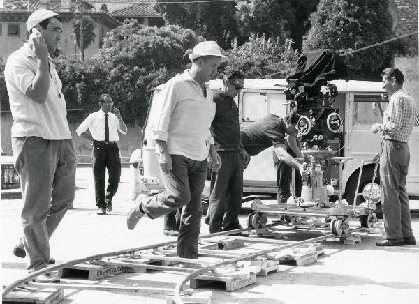 Fotografia sul set di "Frenesia dell'estate" - Zampa, Luigi, 1964 - Al centro, il regista Luigi Zampa cammina in mezzo ai binari della cinepresa mentre a sinistra, Marcello Gatti guarda di fronte. Dietro, operatori non identificati.