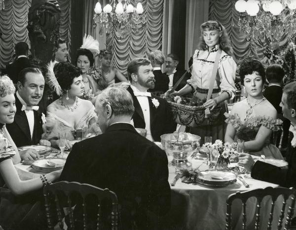 Scena del film "Frou - Frou" - Genina, Augusto, 1955 - Dany Robin, a destra, vestita in abiti da cameriera, con un cesto al collo, incrocia lo sguardo con Gino Cervi seduto al tavolo. Al tavolo, sette attori non identificati.