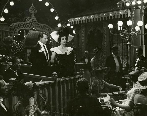 Scena del film "Frou - Frou" - Genina, Augusto, 1955 - Al centro di un locale: a sinistra, Ivan Desny guarda Irene Tunc. L'attrice vestita in abiti eleganti e cappello con piume guarda dritto davanti a sé. Attorno, attori non identificati.