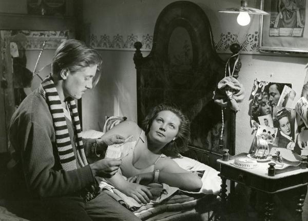 Scena del film "Fuga in Francia" - Soldati, Mario, 1948 - In una stanza, Rosi Mirafiore è sdraiata su un letto in abiti da notte e ha lo sguardo rivolto verso l'alto. Di fianco a lei, a sinistra, Pietro Germi tiene tra le mani un foglio e lo guarda.