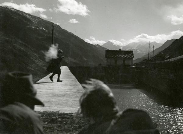 Scena del film "Fuga in Francia" - Soldati, Mario, 1948 - Su una passerella vicino ad un fiume, un attore non identificato, voltandosi, spara ad altri due attori non identificati in primo piano di spalle. Sullo sfondo, i monti.