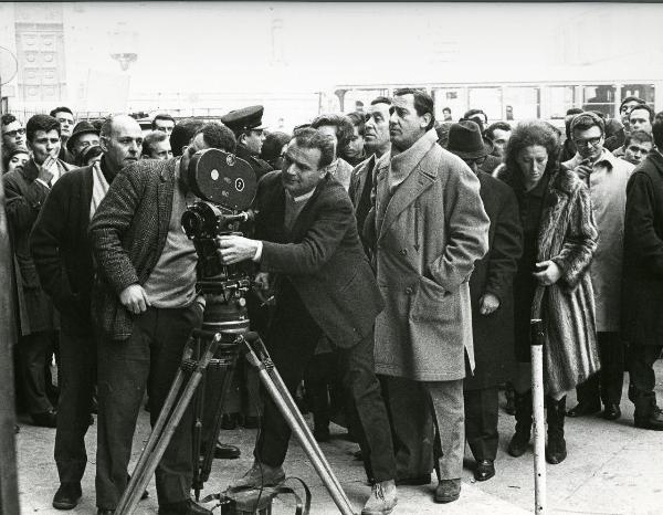 Fotografia sul set di "Fumo di Londra" - Sordi, Alberto, 1966 - In mezzo ad una folla si gira il film "Fumo di Londra". In primo piano, il regista Alberto Sordi mentre guarda la scena che sta riprendendo il macchinista davanti a lui con la cinepresa.