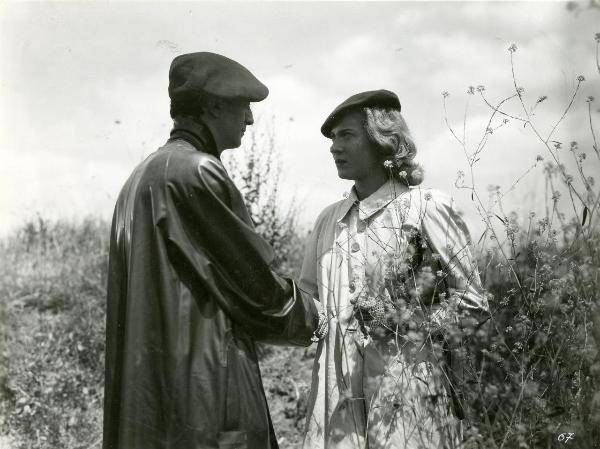 Scena del film "Fumeria d'oppio" - Matarazzo, Raffaello, 1947 - In mezzo a un campo di sterpaglie, Emilio Ghione Jr. a sinistra, dà la mano a Mariella Lotti a destra. I due si guardano negli occhi.