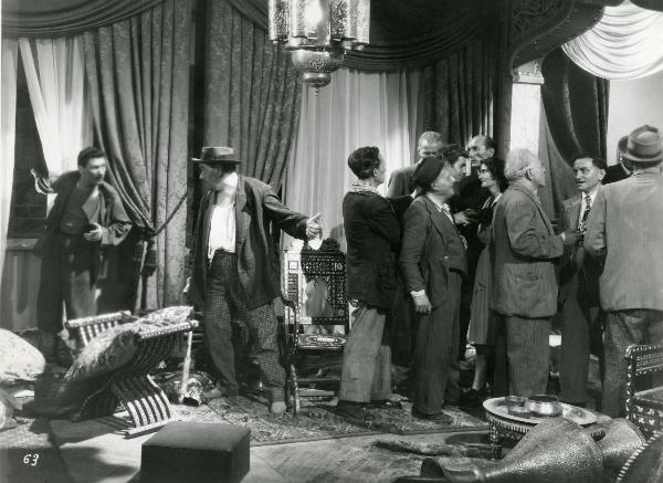 Scena del film "Fumeria d'oppio" - Matarazzo, Raffaello, 1947 - In una sala arredata: a destra, alcuni attori non identificati parlano. A sinistra, due attori non identificati, guardandosi, indicano due zone diverse della stanza.