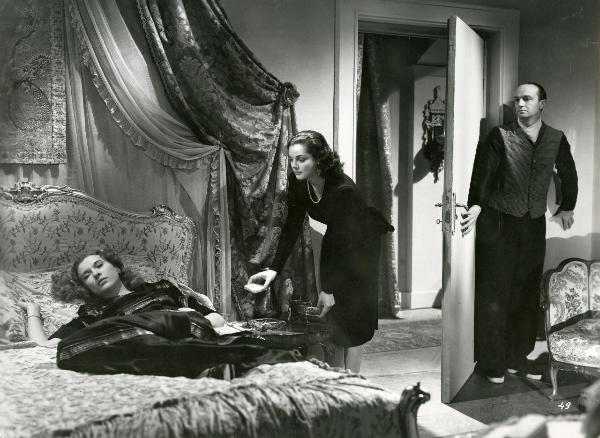 Scena del film "Fumeria d'oppio" - Matarazzo, Raffaello, 1947 - Distesa su un letto, Mariella Lotti. Al centro, un'attrice non identificata si protrae verso di lei. A destra, Emilio Ghione Jr. si nasconde dietro la porta d'ingresso della camera.