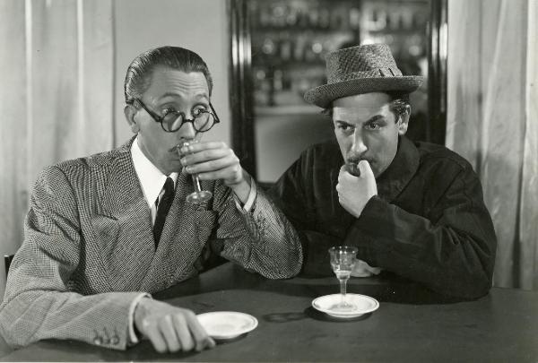 Scena del film "Fuochi d'artificio" - Righelli, Gennaro, 1938 - Seduti a un tavolo: a sinistra, Renato Navarrini che con la mano sinistra si porta il bicchiere alla bocca, a destra, Giuseppe Porelli che si porta la mano sinistra al viso.