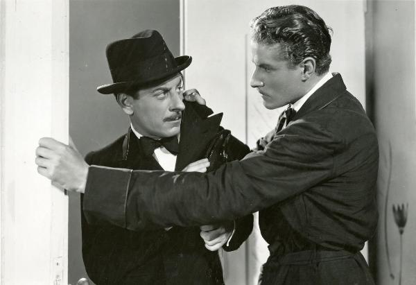 Scena del film "Fuochi d'artificio" - Righelli, Gennaro, 1938 - A destra, Amedeo Nazzari tiene con la mano destra il bavaro della giacca di Giuseppe Porelli mentre la mano sinistra é appoggiata allo stipite di una porta. I due si guardano.