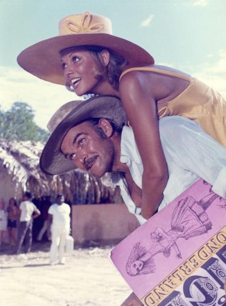 Scena del film "Fuori il malloppo (Popsy Pop)" - Herman, Jean, 1971 - Claudia Cardinale è trasportata in spalla da Stanley Baker. I due, guardando di fronte a loro, sorridono. Lui tiene sotto il braccio sinistro una rivista.