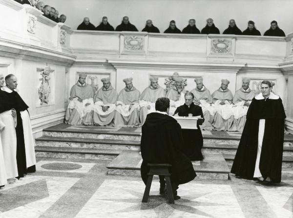 Scena del film "Galileo" - Cavani, Liliana, 1968 - Al centro, un attore di spalle seduto su uno sgabello viene giudicato da un altro attore che gli sta di fronte. Dietro, una schiera di attori in abiti talari di alto rango.