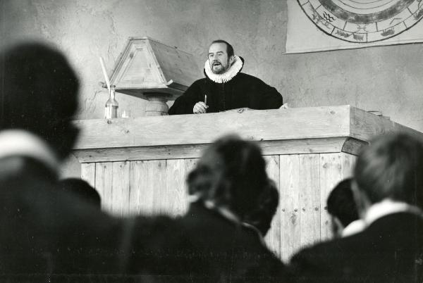 Scena del film "Galileo" - Cavani, Liliana, 1968 - Dall'alto di una cattedra, Cyril Cusack, con una matita in mano, parla ad alcuni attori non identificati di spalle posti in primo piano.