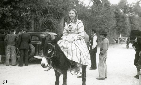 Scena del film "Un garibaldino al convento" - De Sica, Vittorio, 1942 - In sella ad un asino, Carla Del Poggio guarda dritto verso la cinepresa. Dietro, alcuni attori non identificati attorno a un'automobile.