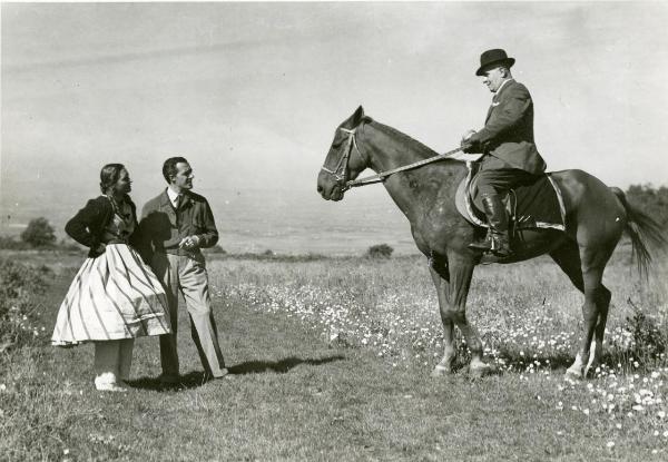 Fotografia sul set di "Un garibaldino al convento" - De Sica, Vittorio, 1942 - In mezzo a un campo: a sinistra, Carla Del Poggio e Vittorio De Sica guardano verso un attore non identificato con cappello in sella a un cavallo, sulla destra.