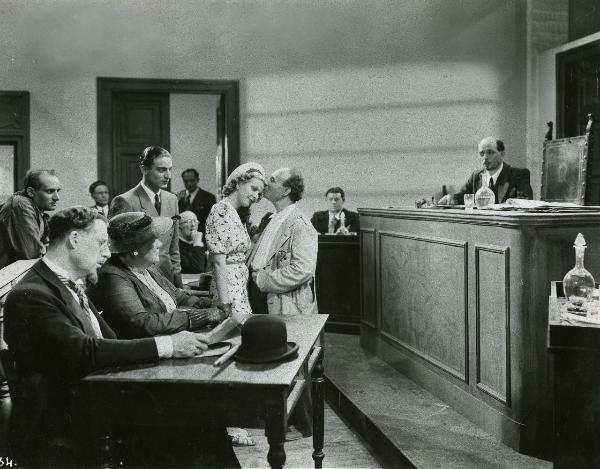 Scena del film "Gatta ci cova" - Righelli, Gennaro, 1937 - In una sala di tribunale: in primo piano seduti ad un tavolo, un attore non identificato e Rosina Anselmi. Al centro, Angelo Musco dà un bacio sulla fronte a Silvana Jachino.