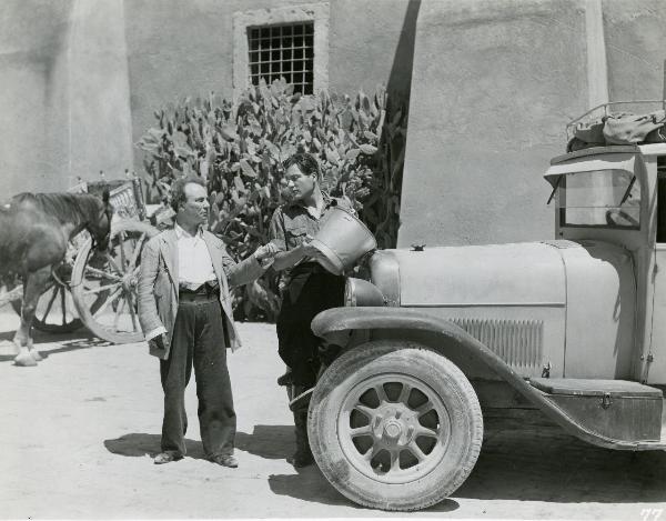 Scena del film "Gatta ci cova" - Righelli, Gennaro, 1937 - Di fronte a un'automobile, Mario Colli, a destra, tiene in mano un secchio e guarda Angelo Musco, a sinistra. Dietro, un carretto con un cavallo e delle piante grasse.