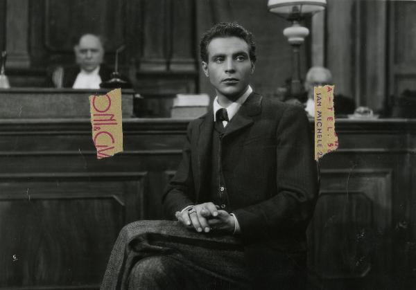 Scena del film "Gelosia" - Poggioli, Ferdinando Maria, 1943 - In un aula: mezza figura di Roldano Lupi seduto a gambe accavallate mentre si tiene la mano destra nella sinistra e guarda davanti a sé. Dietro, due attori non identificati.