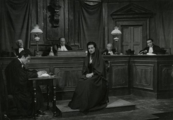 Scena del film "Gelosia" - Poggioli, Ferdinando Maria, 1943 - Al centro, Anna Arena guarda dritto. A sinistra, Roldano Lupi, seduto a uno scrittoio é intento a scrivere. In secondo piano, quattro attori non identificati nei panni di giudici.