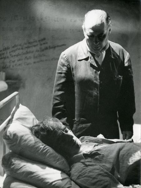 Fotografia sul set di "Il Generale Della Rovere" - Rossellini, Roberto, 1959 - In primo piano, steso a letto e malridotto, Vittorio De Sica. In piedi, dietro di lui il regista Roberto Rossellini lo guarda.