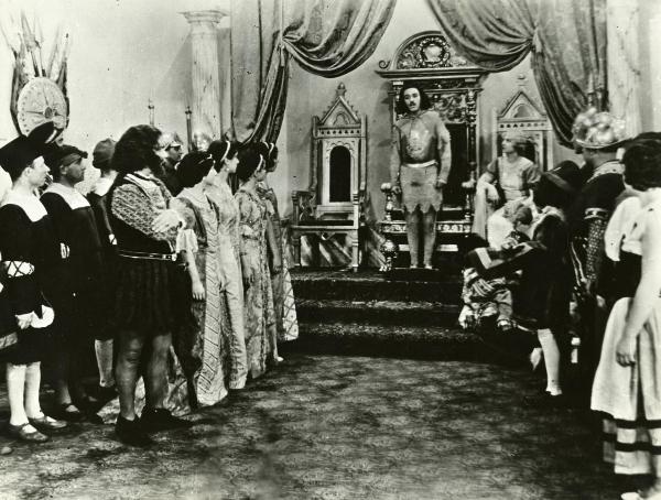 Scena del film "Genoeffa di Brabante" - Zeglio, Primo, 1947 - Nella sala del trono: in secondo piano, un'attore non identificato e Harriet Medin seduta accanto a lui. Attorno a loro, un gruppo di attori non identificati vestiti in abiti da corte.