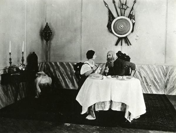 Scena del film "Genoeffa di Brabante" - Zeglio, Primo, 1947 - Seduti a una tavola imbandita: a destra, Aristide Gambini tiene nella mano destra una pipa e guarda intensamente l'attrice non identificata a sinistra che tiene tra le mani una brocca.