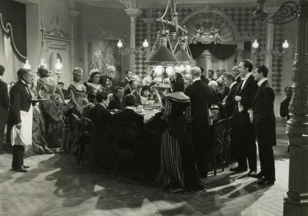 Scena del film "La Gerla di papà Martin" - Bonnard, Mario, 1940 - Attorno a un tavolo da gioco una folla di attori non identificati sono intenti a giocare. A destra: Nino Crisman e Roberto Villa.