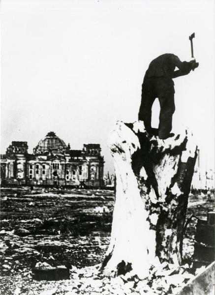 Scena del film "Germania anno zero" - Rosselini, Roberto, 1948 - Un attore non identificato, con in mano un'ascia, è in piedi su un tronco di legno. In secondo piano, un edificio.