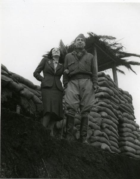 Fotografia sul set di "Giarabub" - Alessandrini, Goffredo, 1942 - Vicino a un fortino costruito con dei sacchi: Doris Duranti è a braccetto con il regista Goffredo Alessandrini. Entrambi, guardando avanti, sorridono.